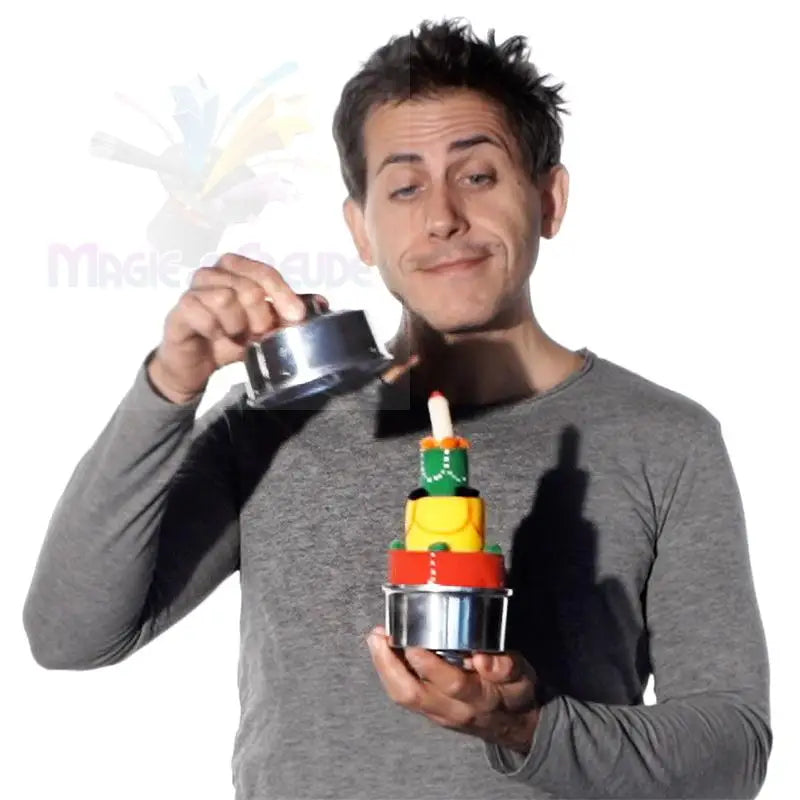 Ein Mann hält einen mehrstöckigen, bunten Kuchen mit einer Kerze, der teilweise von einem Metalldeckel bedeckt ist.