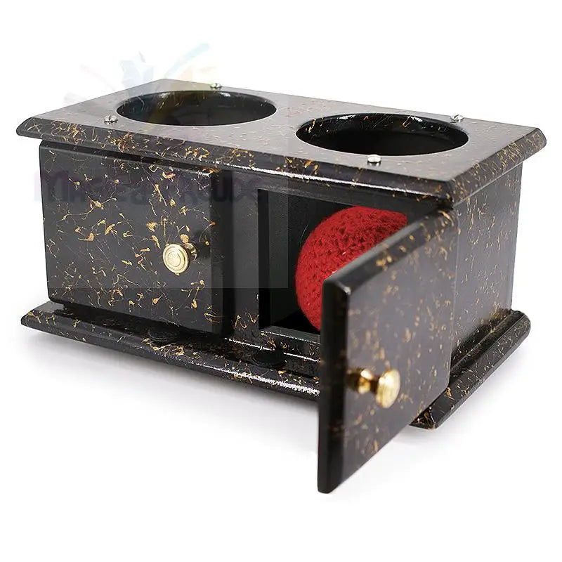 Eine schwarz-gold gesprenkelte Zauberkiste aus Holz mit zwei Löchern im Deckel und zwei Fächern auf der Vorderseite, von denen eines eine rote Kugel enthält.