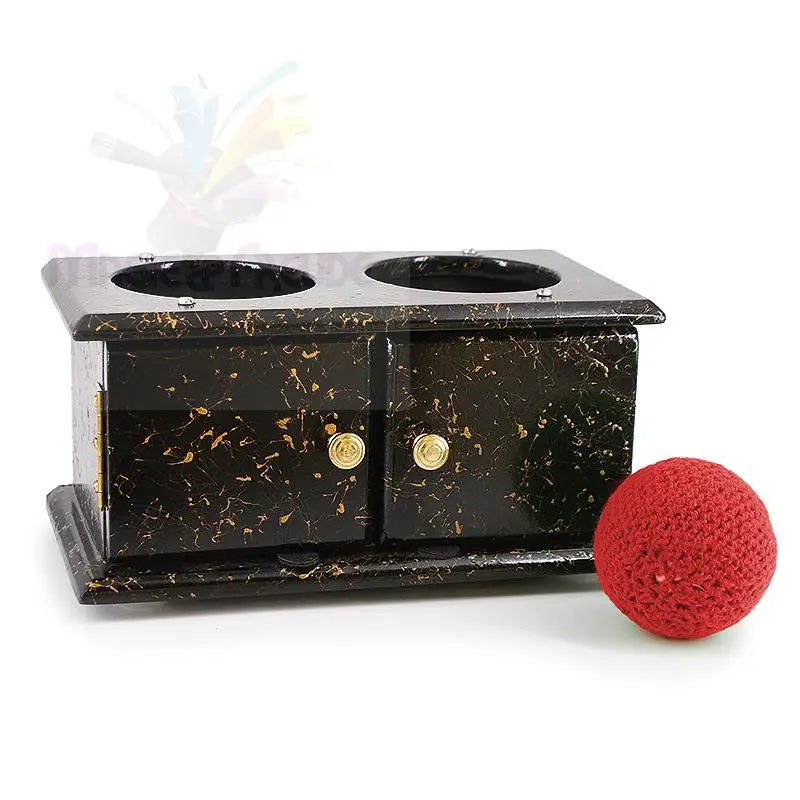 Schwarz-gold gesprenkeltes Zauberrequisit aus Holz mit zwei vertieften Löchern und zwei kleinen Türen, begleitet von einem roten gehäkelten Ball.