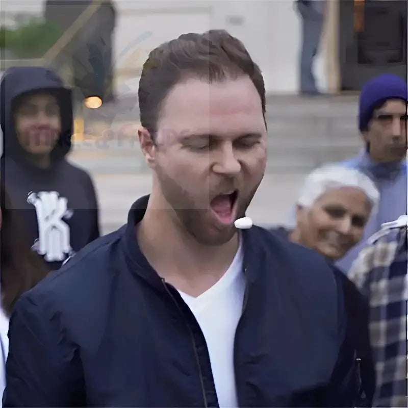 Mann in dunkelblauer Bomberjacke mit weißem T-Shirt darunter, gähnend mit einem weißen Gegenstand im Mund.