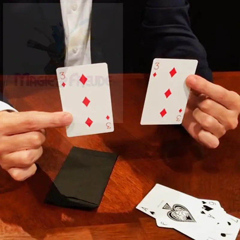 Zwei Hände halten zwei identische Spielkarten hoch, die beide die ’Karo 3’’ offen zeigen, darunter ein gefaltetes schwarzes Tuch und ein Stapel anderer Spielkarten auf einem Holztisch.